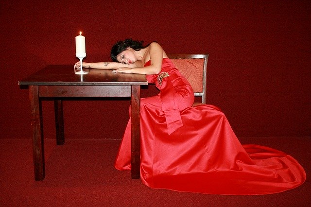 Žena v červených dlouhých šatech s vlečkou, sedící u stolu se zapálenou svící